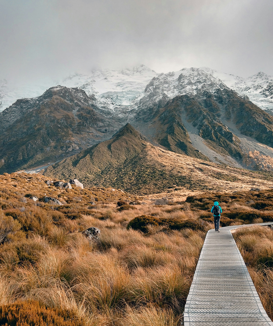 A backpacker walks along a wooden trail towards snow-covered mountains in the New Zealand landscape.